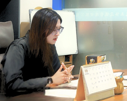 The image shows a woman named YUKA sitting at a desk. She is wearing a black long-sleeved shirt and has long dark hair. The desk has several items on it, including a calendar, a small decorative item, and some papers. In the background, there is a computer monitor and a glass partition with some text.